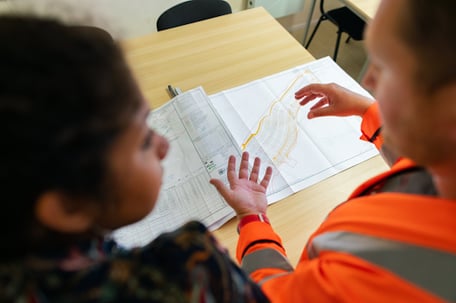 Two people are reviewing building documents. The documents are on white paper and set on top of a wooden table. One person is wearing a bright orange shirt.