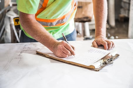 This person is reviewing a building document on a brown clipboard. They are wearing a bright yellow safety vest and have a tape measure on their hip.