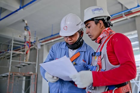 Two people are reviewing a document. They are both wearing white hard hats and gloves. One person is wearing a red long sleeve shirt, and the other is wearing a blue button-down shirt.
