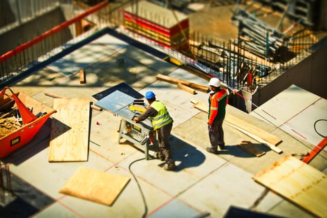 Two construction workers, both wearing safety vests and hard hats, are on a  job site.