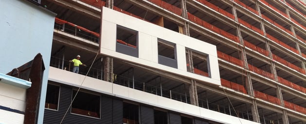 A construction worker in a yellow shirt standing on the second story of a job site. He is directing the installation of a suspended EIFS panel.