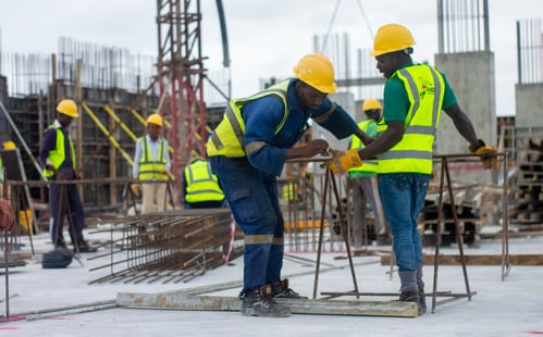 Construction workers wearing yellow hard hats and yellow safety vests are conversing while on a job site.